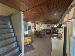 Un interior de varios niveles con una escalera, una sala de estar con sofá y sillones, un televisor y una cocina con refrigerador.