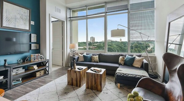 Ein modernes Wohnzimmer mit großem Fenster, einer Couch, zwei Holztischen und einem Fernseher auf einem Regal.