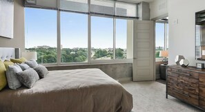 Ein Schlafzimmer mit großem Fenster, einem Bett, einer Kommode und einer Tür.