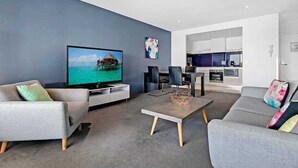 Un saló modern amb televisió plana, sofàs grisos, una taula de centre i una cuina al fons.