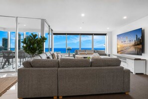Un salon moderne avec un grand canapé gris, un coin repas avec des chaises, une grande fenêtre donnant sur l’océan et un tableau accroché au mur.
