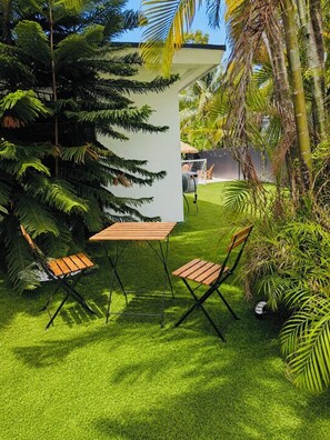 A patio with a table and two chairs, surrounded by lush greenery and a white structure.