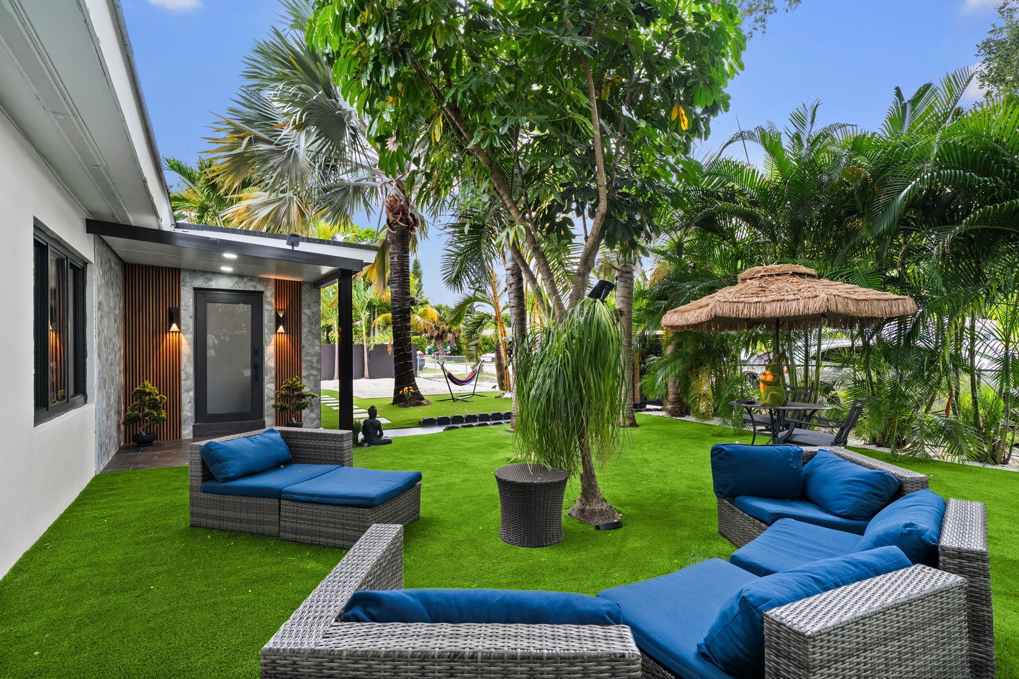 A modern outdoor patio with wicker furniture, a thatched umbrella, and lush greenery.