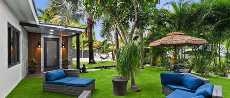 A modern outdoor patio with wicker furniture, a thatched umbrella, and lush greenery.