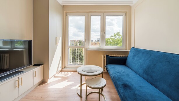 En moderne stue med en blå sofa, et rundt sofabord, et fjernsyn på et skab og store vinduer med udsigt over byen.