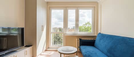 A modern living room with a blue sofa, a small round table, a TV on a cabinet, and large windows with a city view.