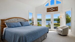 Un dormitorio con una cama grande, una silla y vista a un lago a través de amplias ventanas.