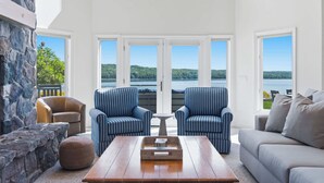 Amplia sala de estar con una pared de acento de piedra, una mesa de centro de madera, dos sillones a rayas, un sofá color beige y ventanas grandes con vistas a un lago y colinas.