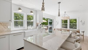 A modern kitchen with white cabinetry, a large island, pendant lighting, and large windows with a view of greenery. - Luxury Hamptons Retreat, Heated Pool + Firepits (Water Mill)