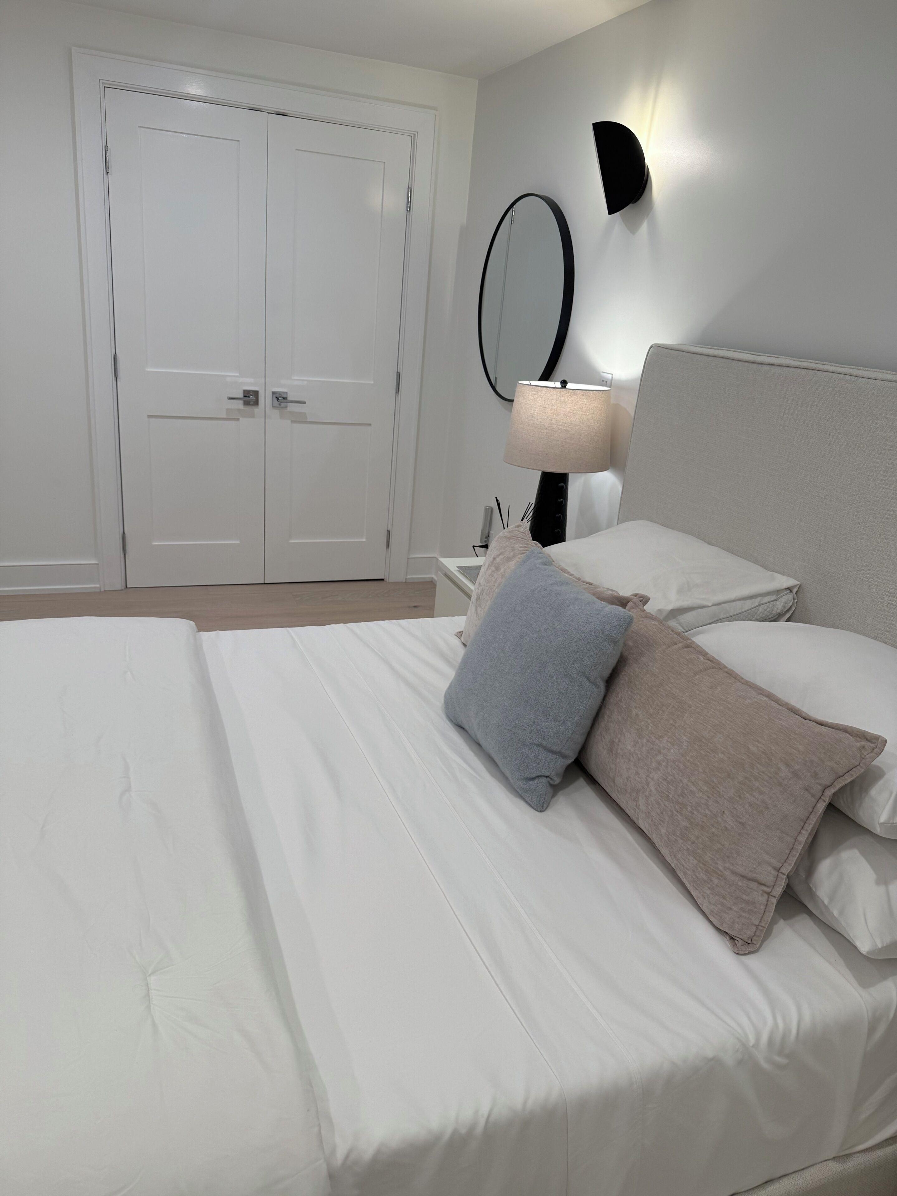 A bedroom with a bed, pillows, a nightstand, a lamp, a mirror, and a door.