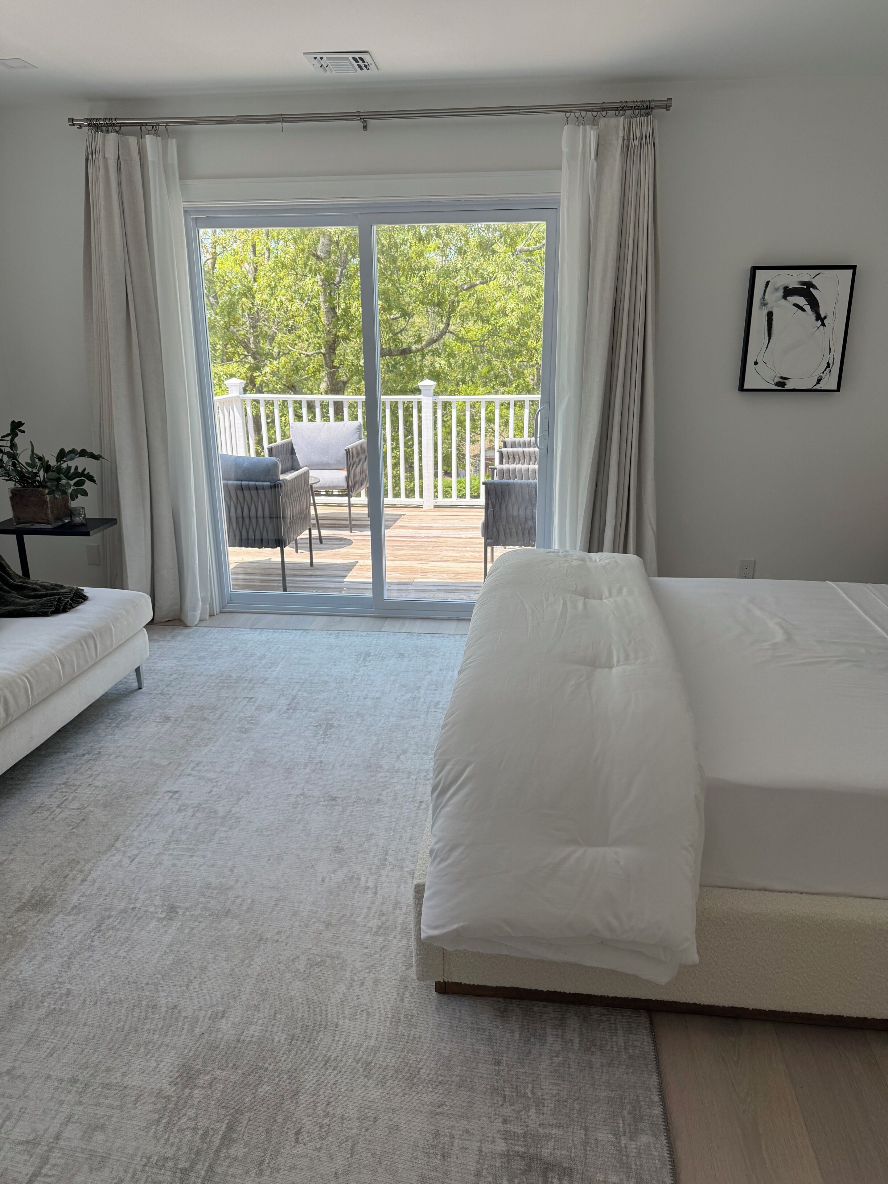 A bedroom with a large bed, a sofa, a small table with a plant, and a view of a balcony with outdoor furniture.