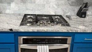 Une cuisine avec des armoires bleues, une hotte de cuisinière en acier inoxydable, un plan de travail en granit, une cuisinière à gaz, un four encastré et un bloc à couteaux.