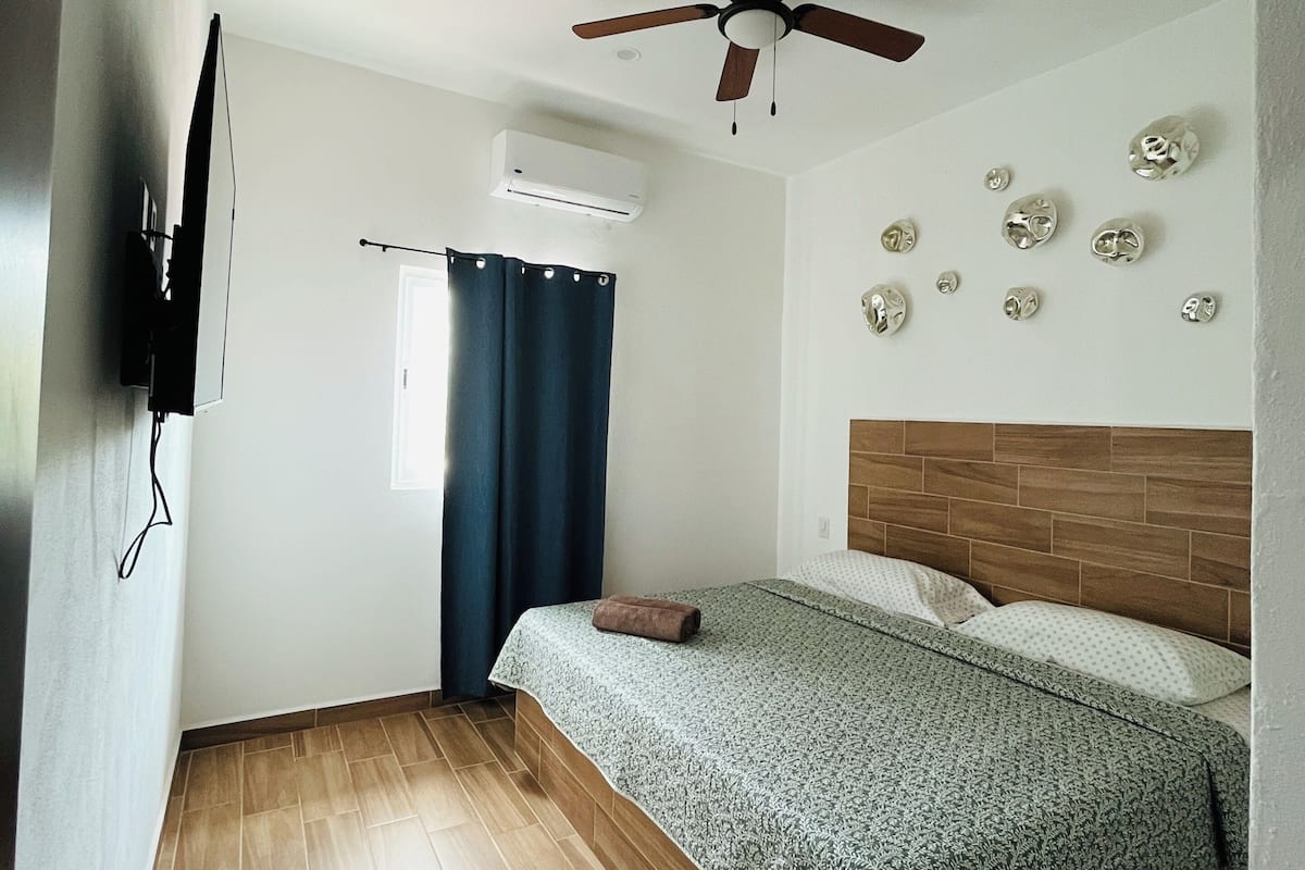 A bedroom with a bed, a ceiling fan, an air conditioner, a window with curtains, and a wall with decorative plates.
