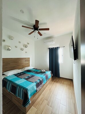 Une chambre à coucher avec une tête de lit en bois, un lit recouvert d’une couvre-lit à motifs, un ventilateur de plafond, un appareil de climatisation, une fenêtre avec des rideaux et un téléviseur fixé au mur.