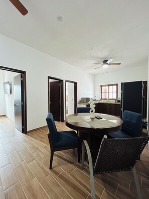 Una habitación amplia con una mesa de comedor redonda, dos sillas azules, un ventilador de techo y una cocina al fondo.