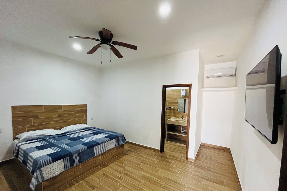 A bedroom with a wooden headboard, a bed with a blue and white checkered bedspread, a ceiling fan, and a wooden floor.