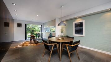 A modern dining area with a round table, chairs, a pendant light, and a view of the kitchen and garden.