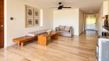 Una sala moderna con un sofá, un banco de madera y una mesa de centro. Hay un ventilador de techo y un refrigerador en la habitación.