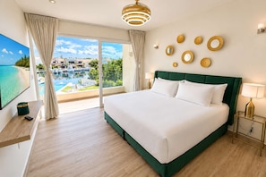 A hotel room with a large bed, a TV, a desk, and a view of a swimming pool and boats. - 3BR · Marina-view terrace · Pool (Puerto Aventuras)