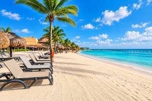 A beach with rows of sun loungers, palm trees, and thatched umbrellas. - 3BR · Marina-view terrace · Pool (Puerto Aventuras)