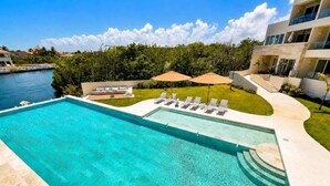 Une piscine avec vue sur un plan d’eau, un espace verdoyant avec des chaises longues et un bâtiment moderne doté de balcons.