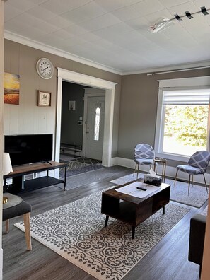 A living room with a TV, two chairs, a coffee table, patterned rugs, a clock, and a door leading to another room. - Spacious house near White Water Walk and many attractions (Niagara Falls)