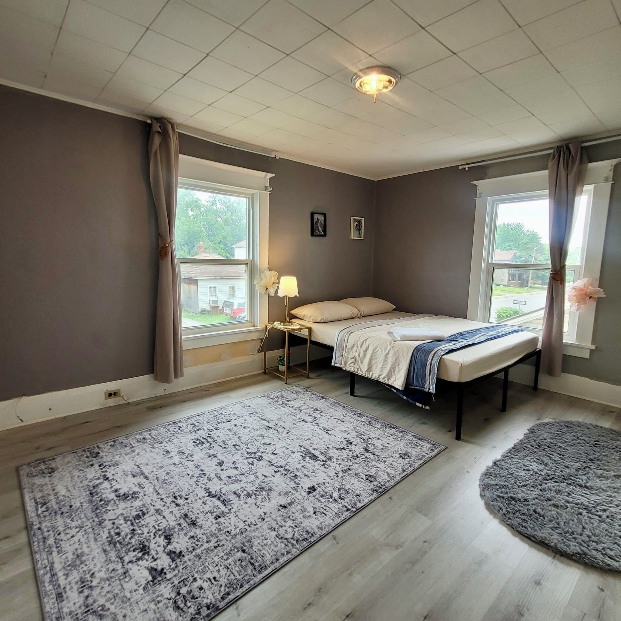A bedroom with a bed, two windows, a nightstand, a lamp, and a rug.