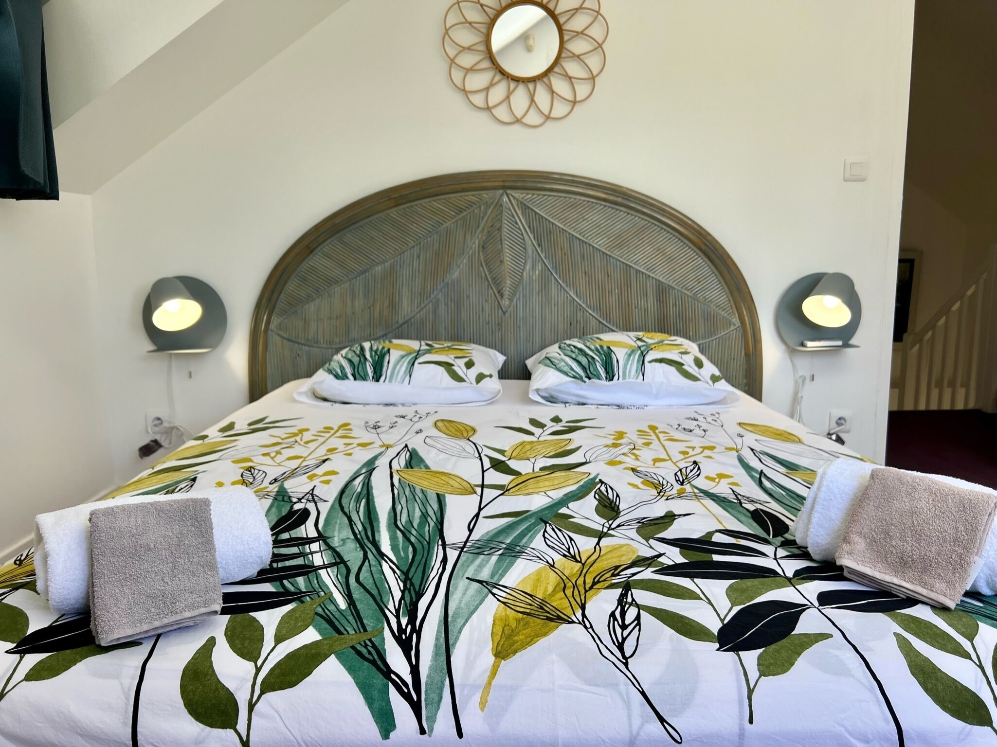 A bed with a leaf-patterned bedspread, two bedside lamps, and a round mirror on the wall.