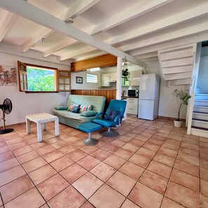 Ein Wohnzimmer mit einer Couch, einem Sessel, einem kleinen Tisch, einem Ventilator und einer Küchenzeile mit Kühlschrank und Mikrowelle.