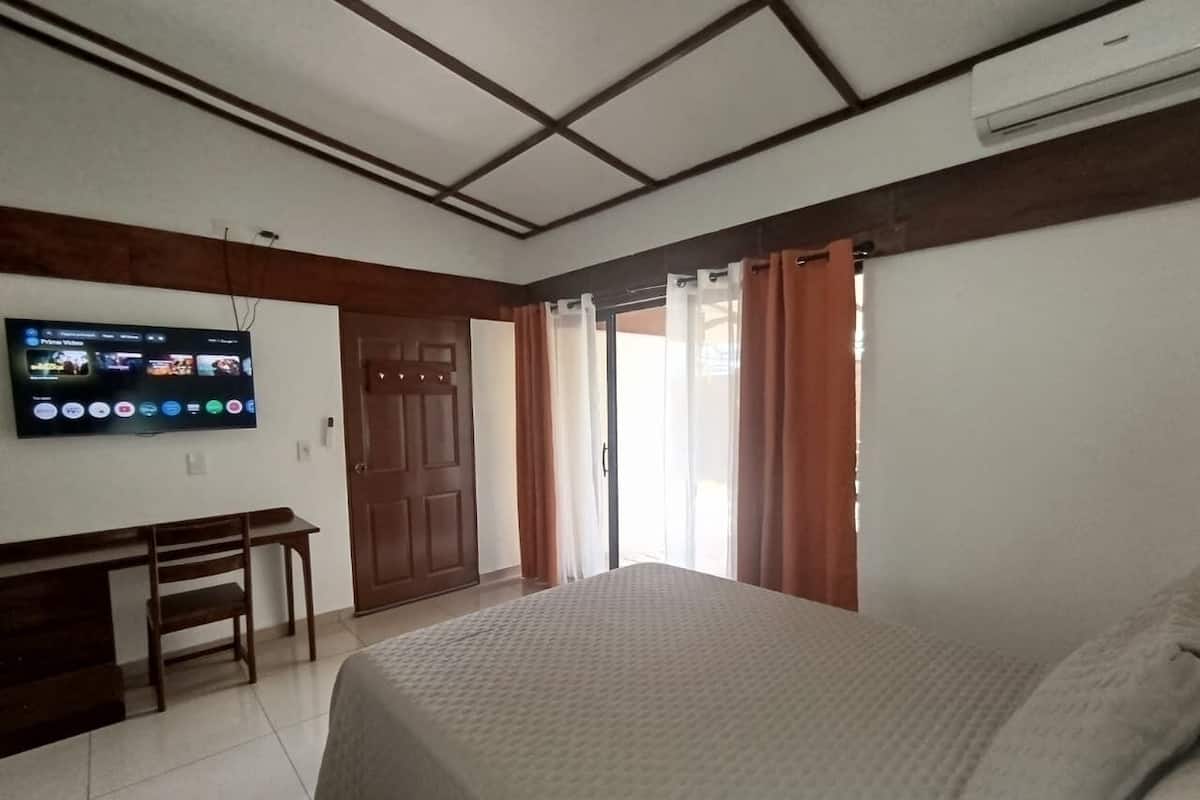 Un dormitorio con cama, escritorio, televisor montado en la pared y puerta corrediza que da a un balcón.