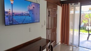 Habitación con una televisión de pantalla plana grande montada en la pared, una mesa de madera, una puerta con panel de vidrio y vistas al exterior.