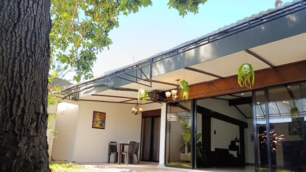 Un edificio moderno con un área exterior cubierta, una mesa de comedor, sillas y plantas colgantes.