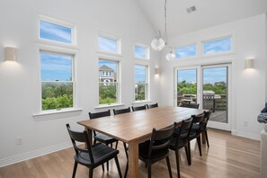 A dining room with a wooden table and chairs, large windows, and a view of the outdoors. - New remodel 2026 - walk to beach, heated pool, hot tub, dogs ok (Nags Head)
