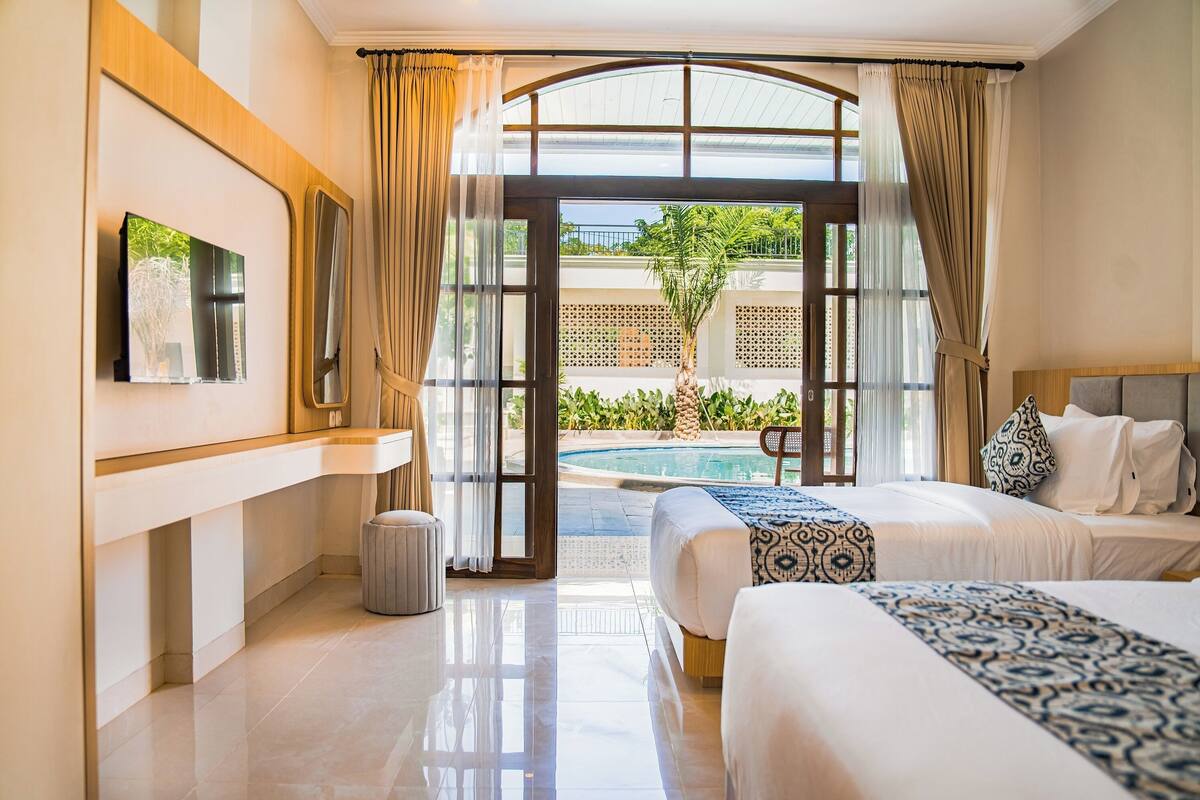 Habitación de hotel con cama, televisor, un basurero pequeño y vista a una piscina y zonas verdes.