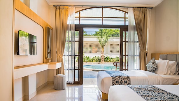 A hotel room with a bed, a TV, a small trash bin, and a view of a pool and greenery.
