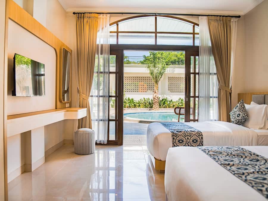 Habitación de hotel con cama, televisor, un basurero pequeño y vista a una piscina y zonas verdes.