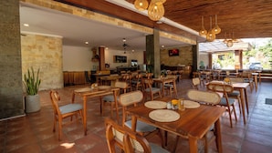 A spacious outdoor dining area with wooden tables and chairs, a stone wall, and a ceiling with woven light fixtures.