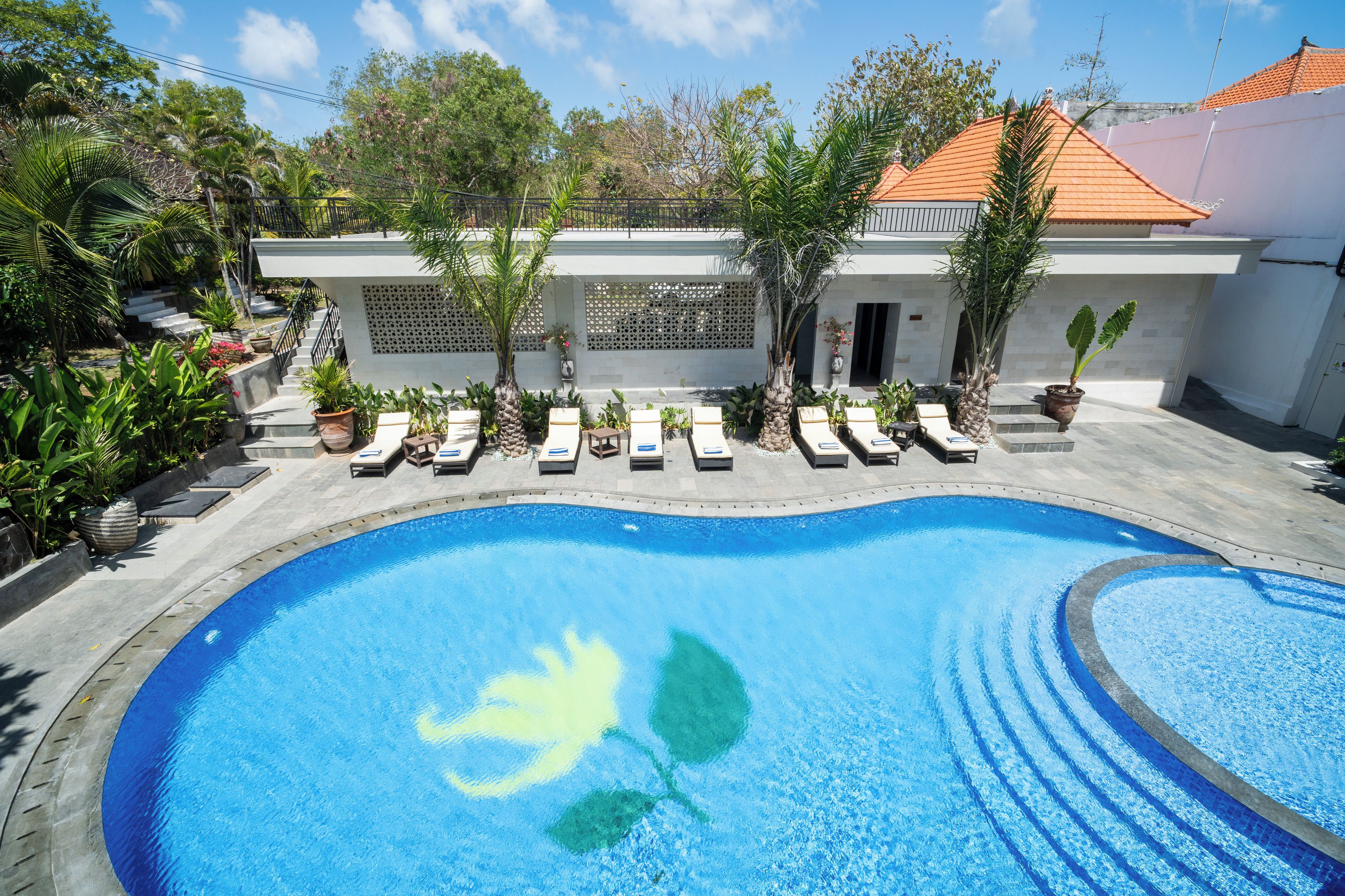 A swimming pool with a sunbather and lounge chairs, surrounded by potted plants and a building with a tiled roof.