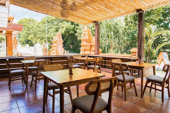 A restaurant with wooden tables and chairs, a thatched roof, and a view of temple structures.