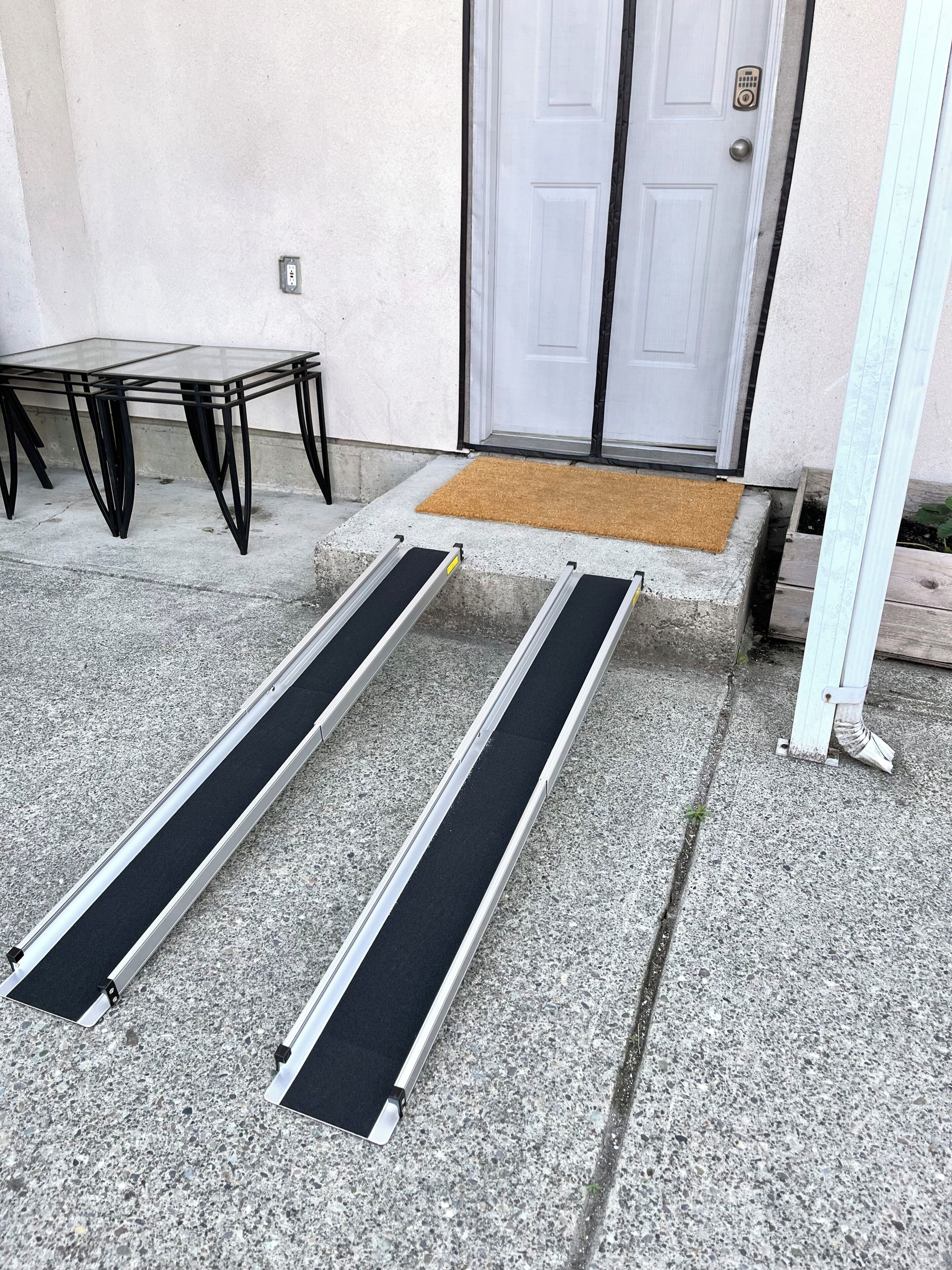 En støbt veranda med et glasbord og metalstole, en hvid dør og en dørmåtte.