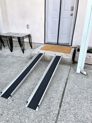 Un porche en béton avec une table en verre et des chaises en métal, une porte blanche et un paillasson.