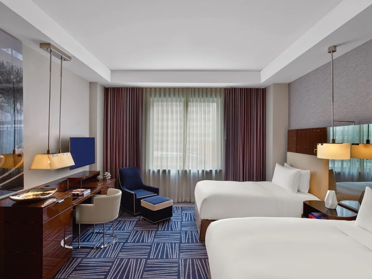Ein Hotelzimmer mit Schreibtisch, zwei Betten, einem Sessel und einem großen Fenster mit Vorhängen.