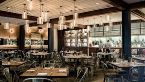 Interno di un ristorante con diversi tavoli e sedie, lampade a sospensione e una zona cucina visibile.