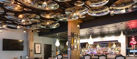 Zona de bar con varios espejos colgantes, barra con botellas y asientos con mesas y sillas.