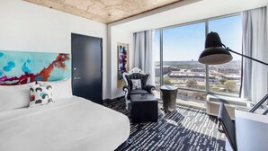 A hotel room with a large bed, a desk, a chair, a window with a city view, and a ceiling with exposed beams.