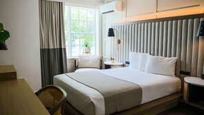 A hotel room with a large bed, a desk, a chair, a small table with a lamp, and a window with curtains.