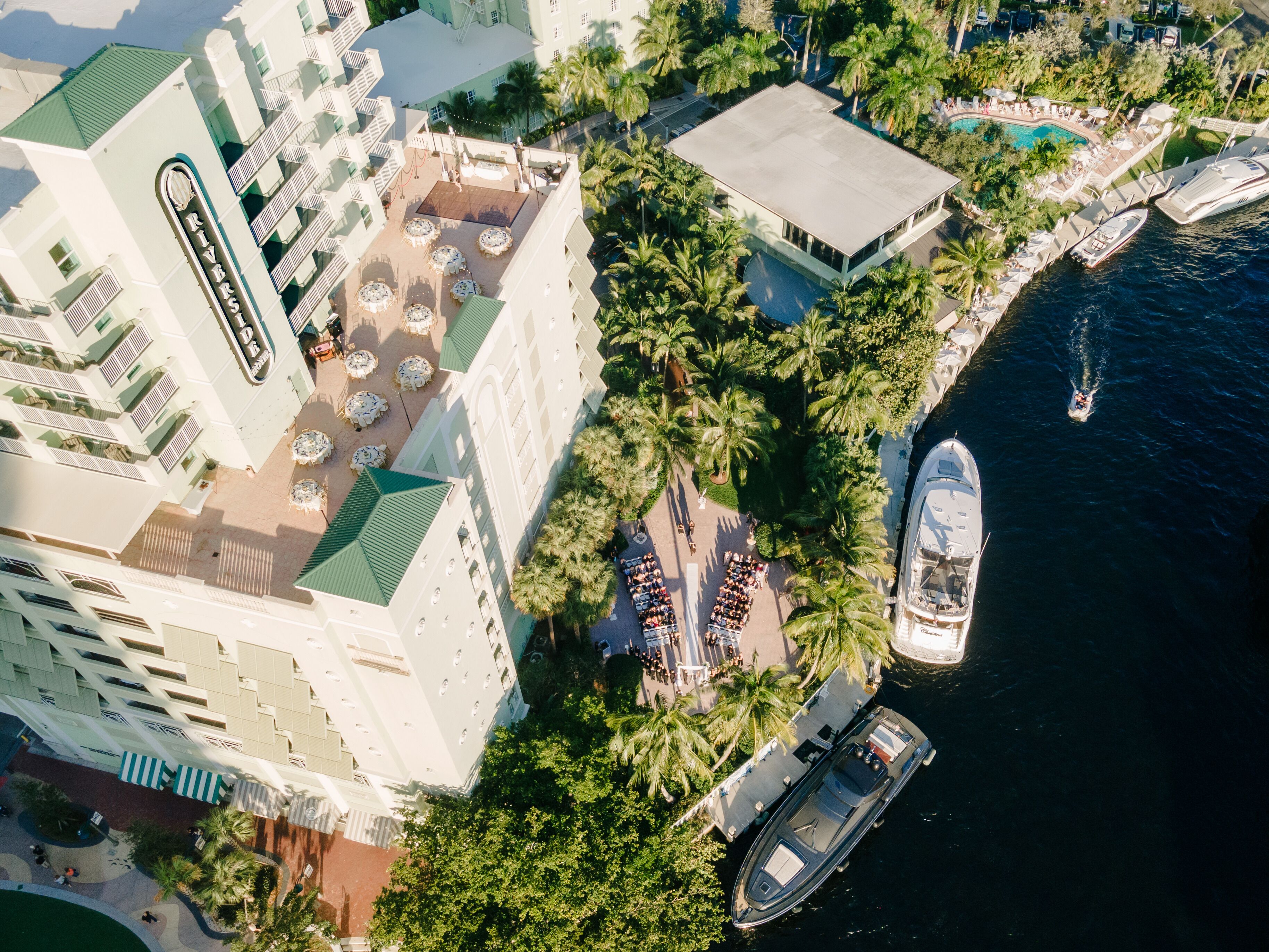 An aerial view of a hotel with a rooftop pool, palm trees, and a marina with boats.
