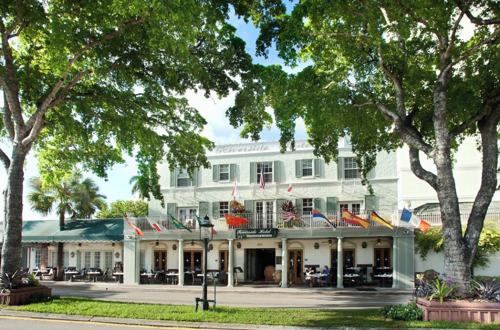 Ein Hotel mit überdachter Außenbestuhlung, mehreren Flaggen und einem großen Baum im Vordergrund.