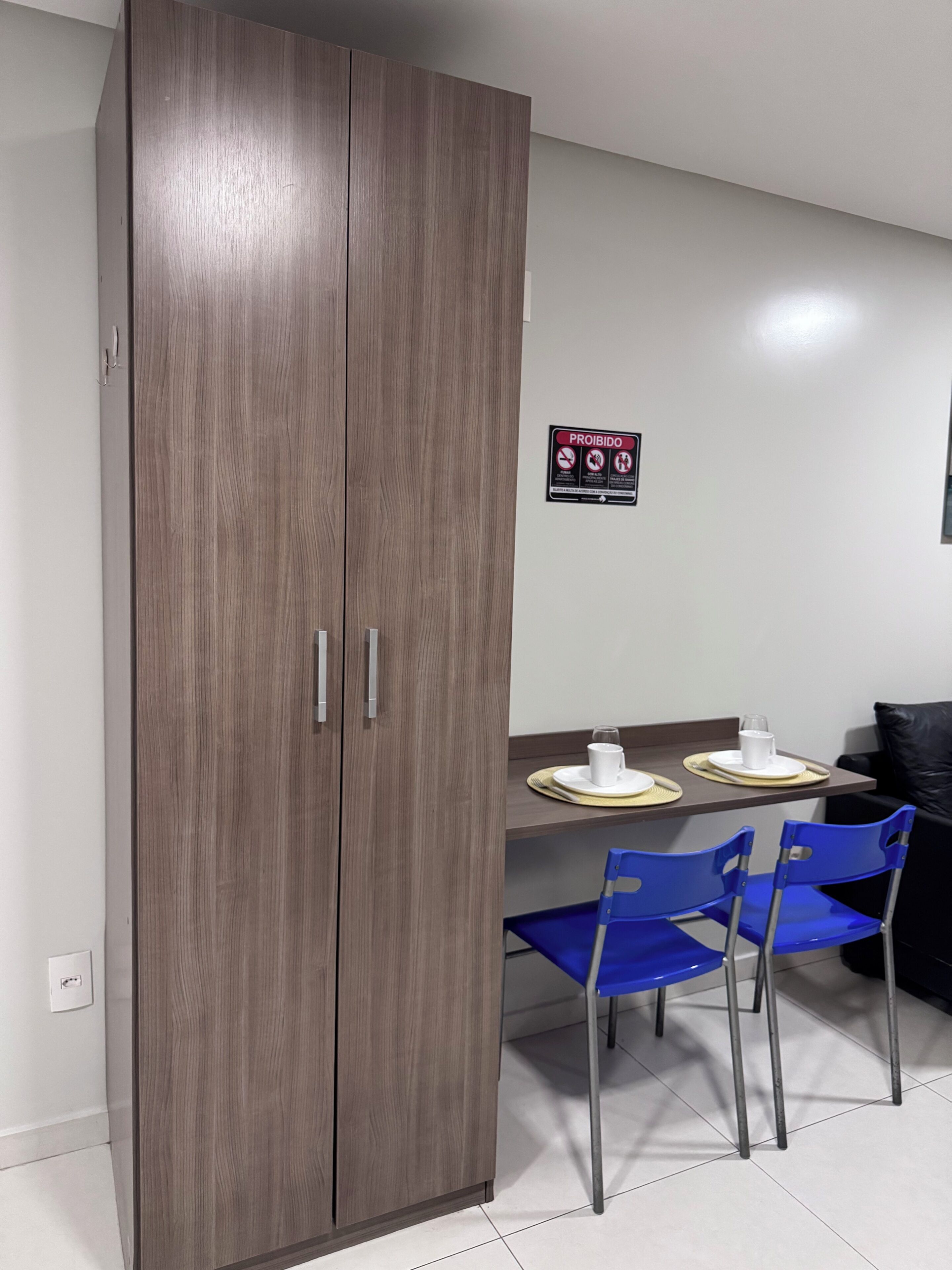 A room with a wooden wardrobe, a dining table with chairs, and a 'PROHIBIDO' sign on the wall.