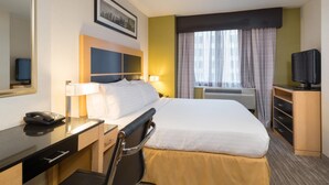 Habitación de hotel con cama, escritorio, silla, lámpara, televisor y un cuadro con una vista urbana.